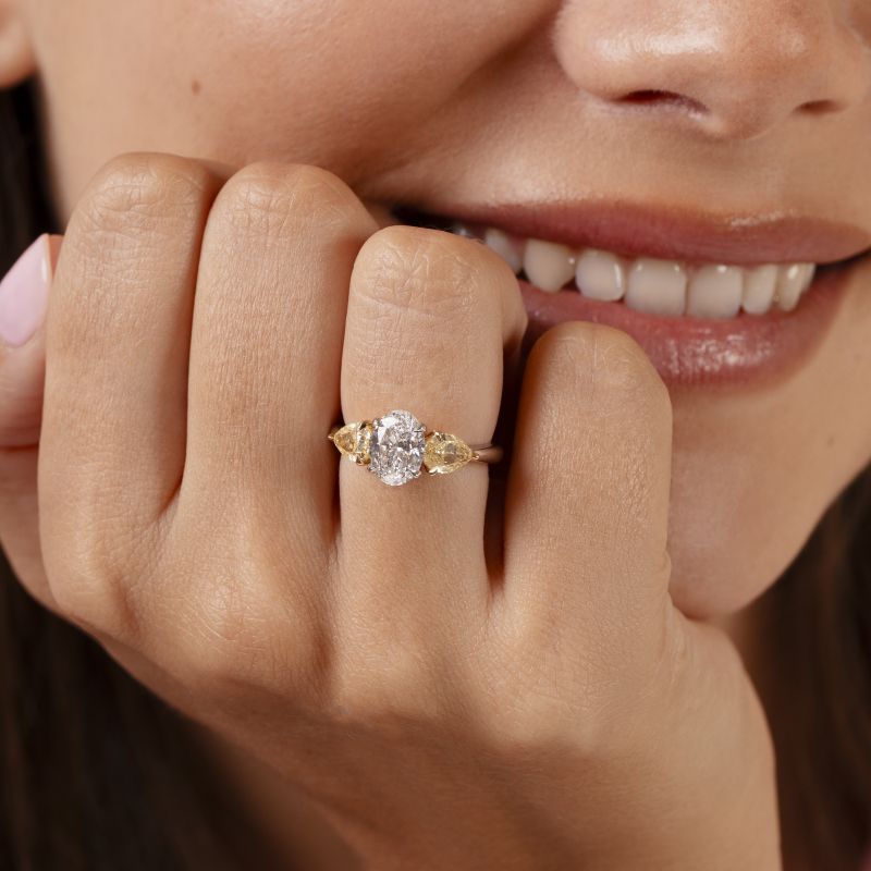 Three stone diamond engagement ring worn on a model's hand, featuring an oval cut white diamond centre stone with fancy yellow pear shaped diamond side stones on a platinum band.